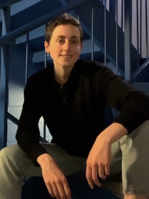 A portrait of Sophie, a white person with very short brown hair. She is sitting in a relaxed position and smiling at the camera.